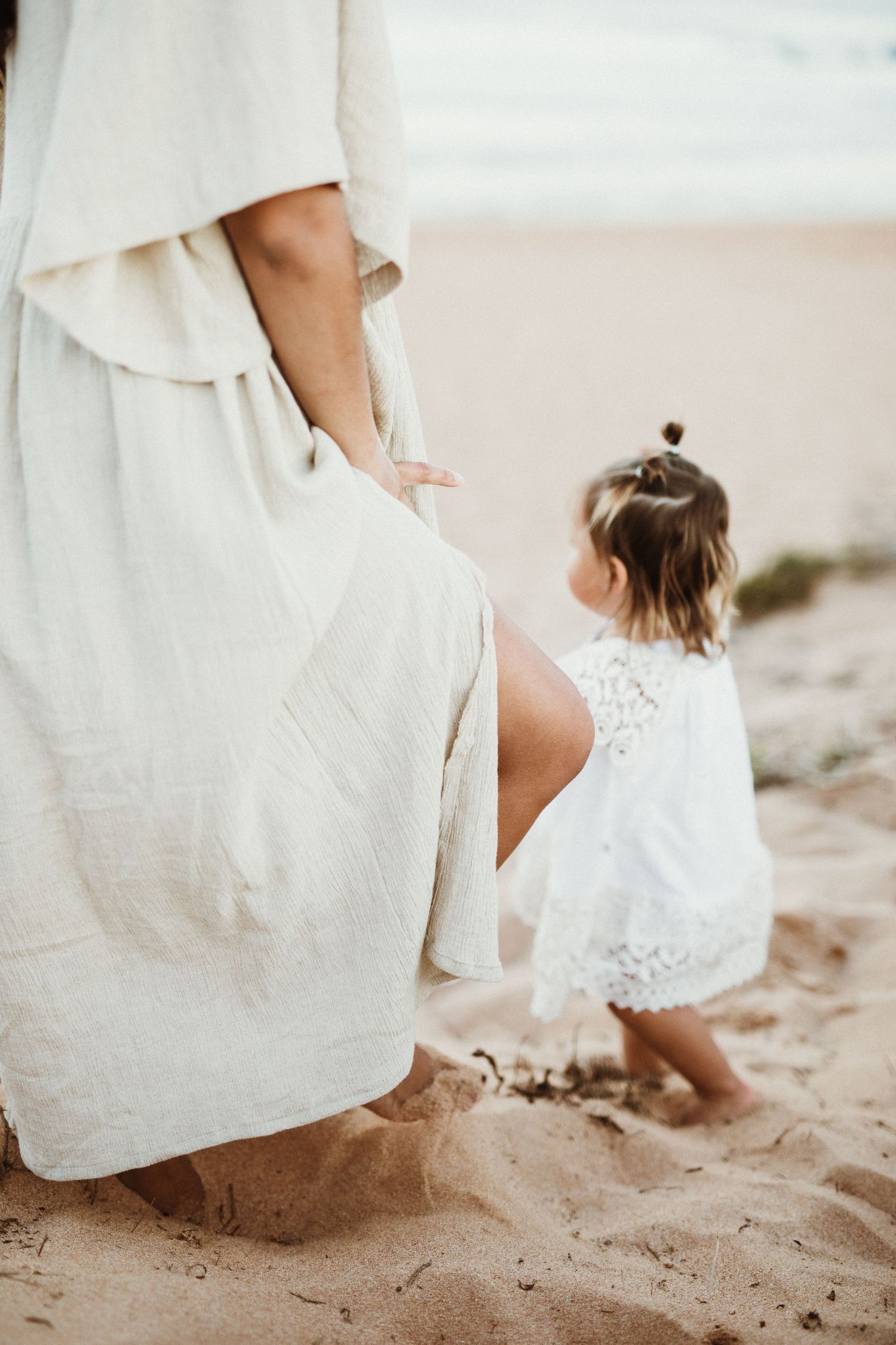A toddler in a white lace dress stands beside her mother on a sandy beach.