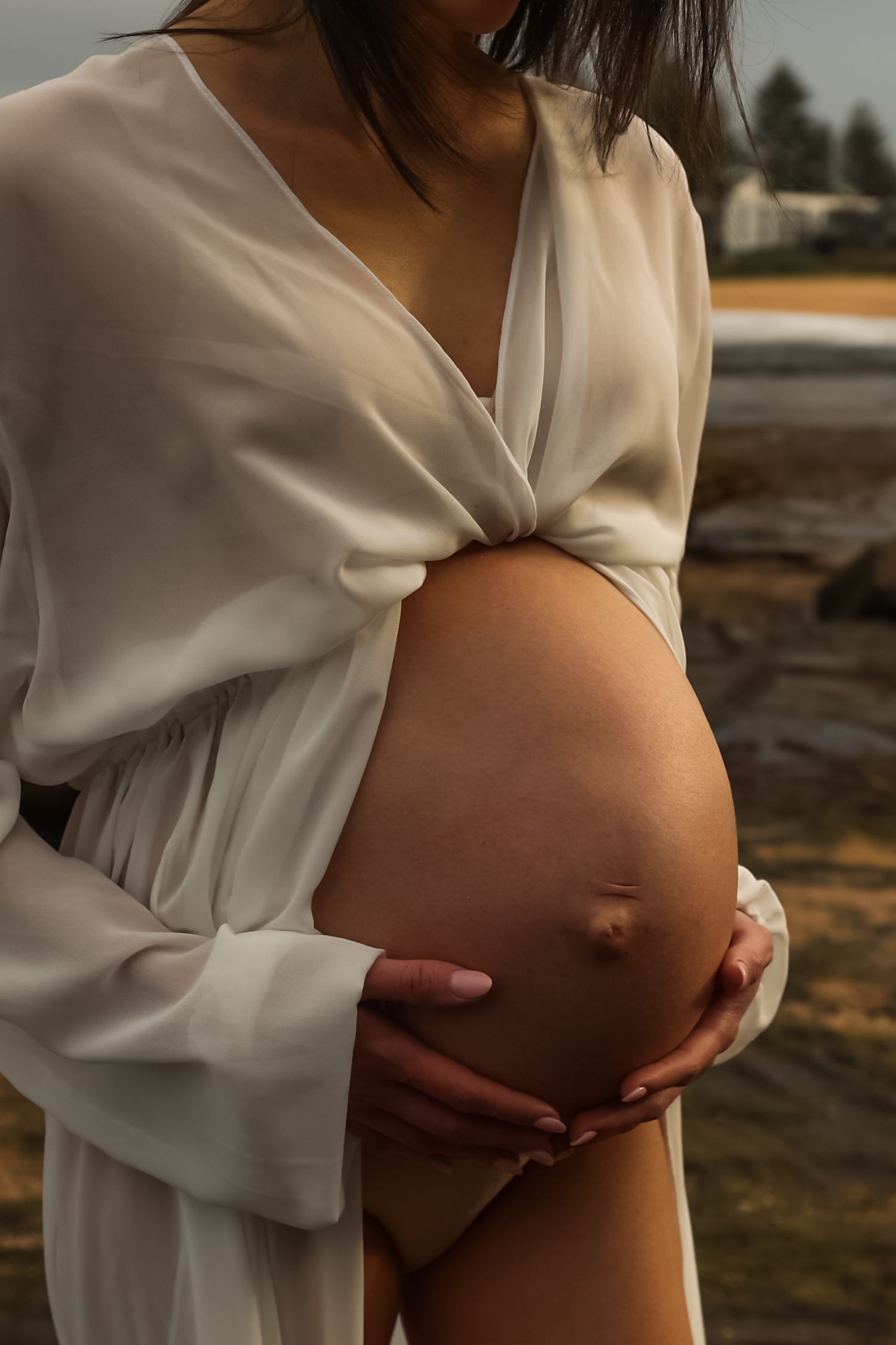 Close-up of a pregnant woman in a white wrap dress cradling her baby bump near a rocky beach.