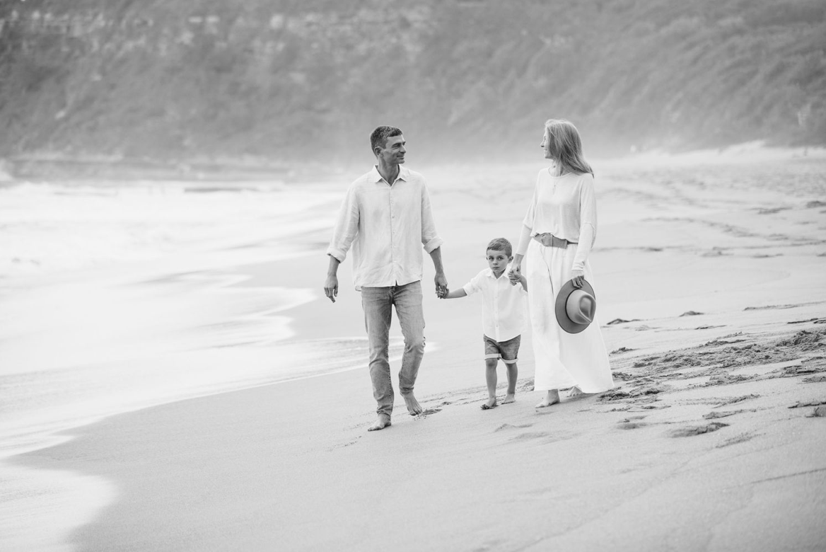 A family of three walks hand in hand along a beach shoreline in a black and white photo.