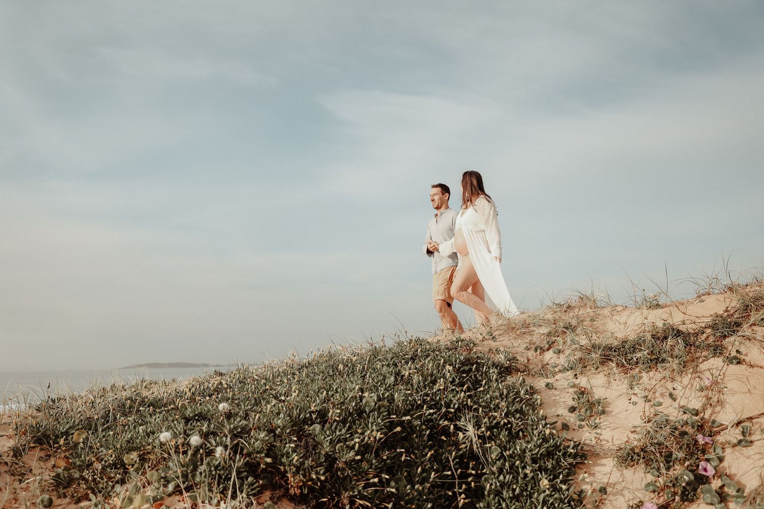Couple in white outfits walking hand in hand on sandy coastal dune with wildflowers and ocean view.