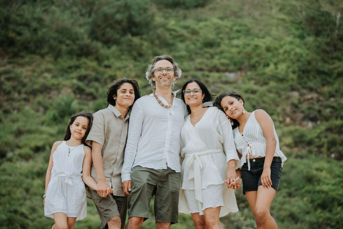 A joyful family of five poses together outdoors surrounded by lush green hills.