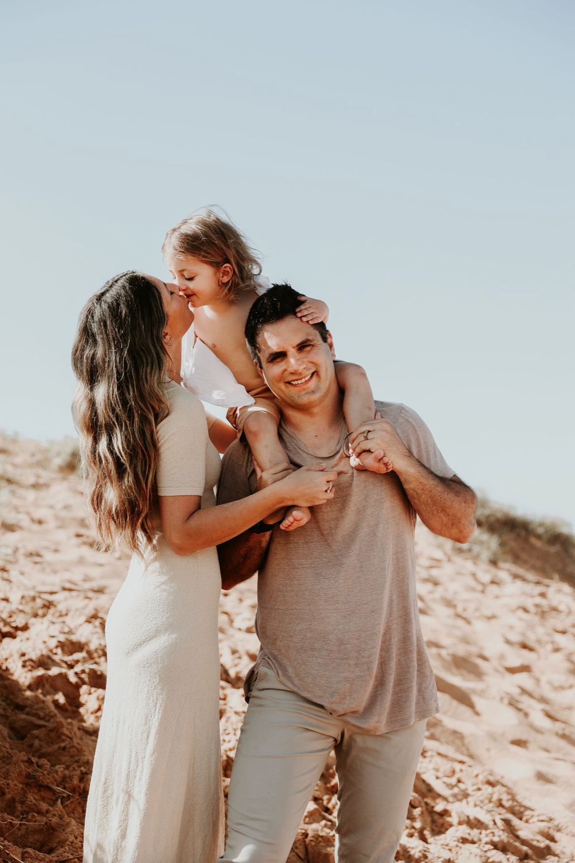 A smiling couple at the beach with their young daughter sitting on the mans shoulders.