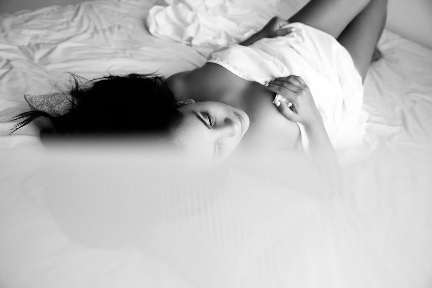 Black and white photo of dark-haired woman lying on white bed sheets in soft natural light.
