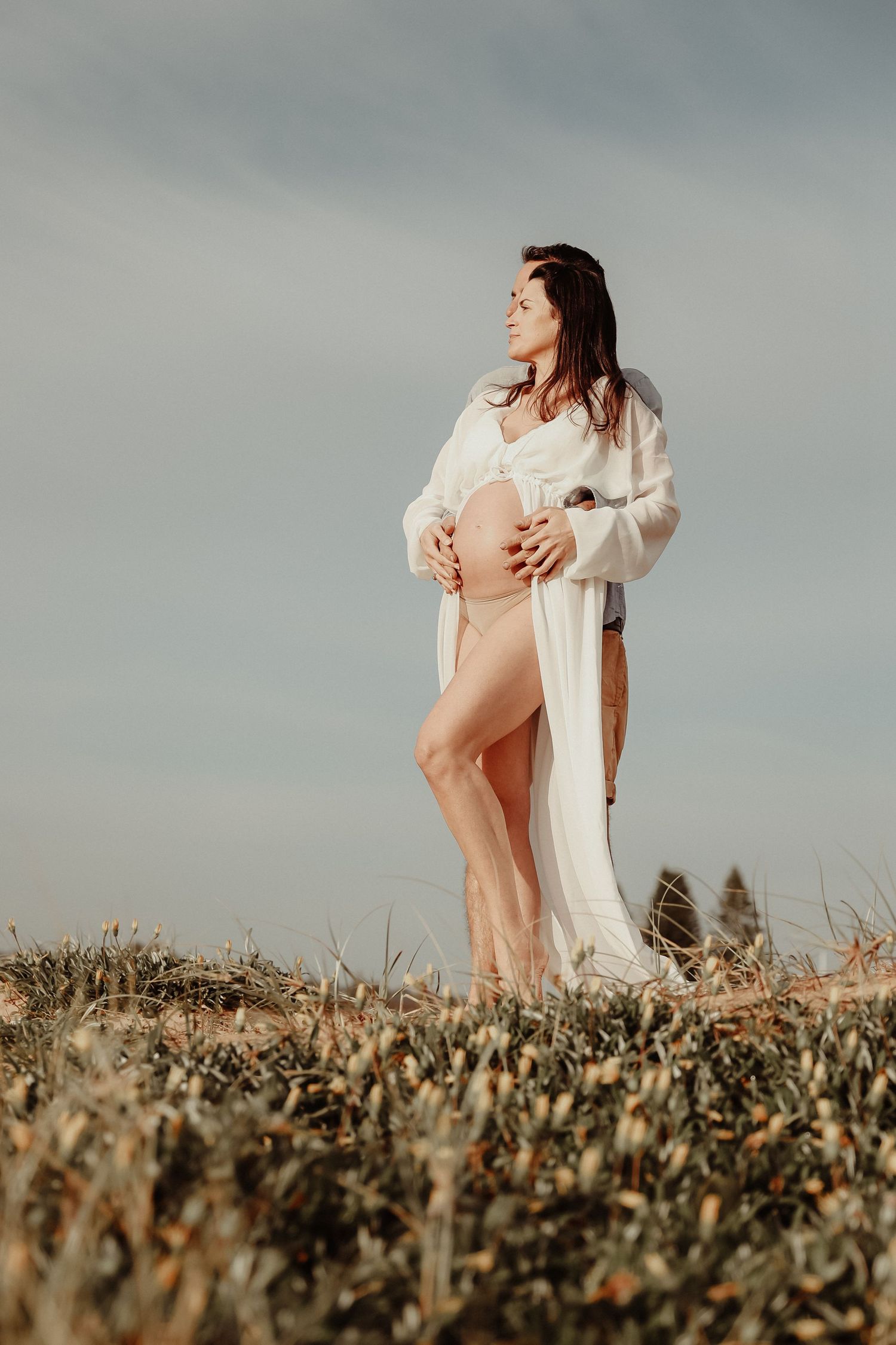 Pregnant woman in white robe holding baby bump outdoors in a field under cloudy sky.