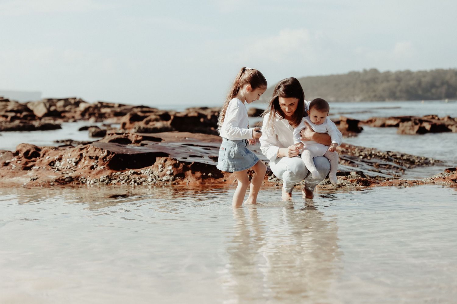 Mother crouching with two young children exploring shallow rocky beach tide pools.