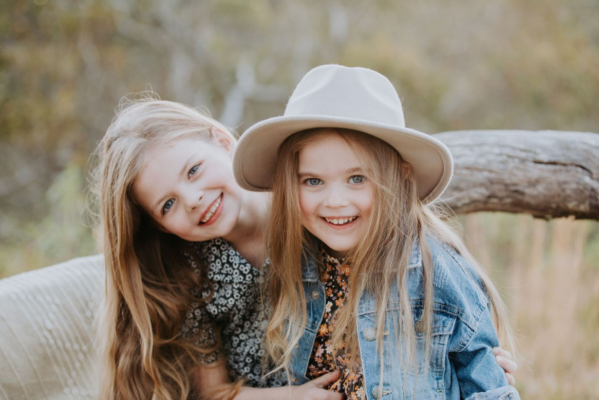 Two smiling blonde sisters pose together outdoors, one wearing a white hat and denim jacket.