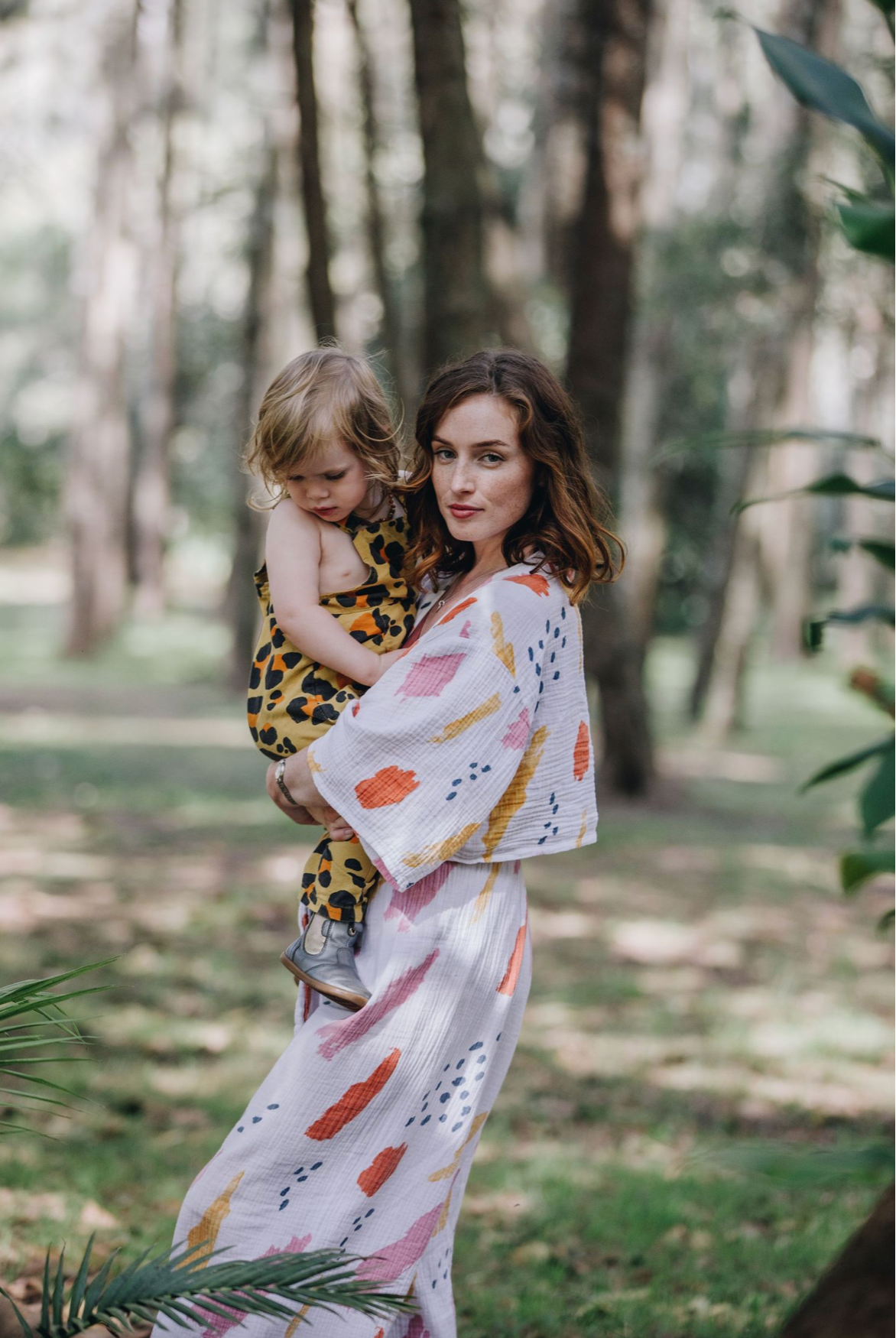 Elegant mother in colorful printed kimono holding her toddler in a sunlit forest setting.