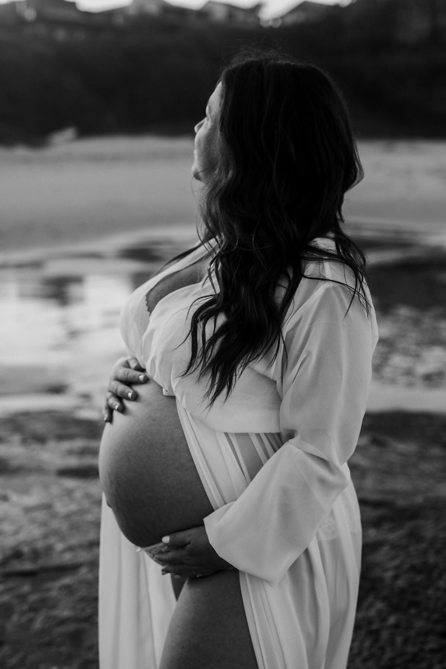 Black and white side profile of pregnant woman in white robe cradling bare baby bump near water.