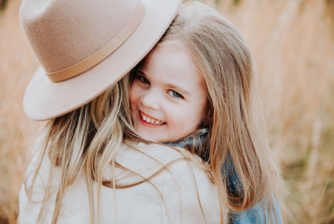 A smiling young girl with long blonde hair wears a beige hat in a warm outdoor setting.
