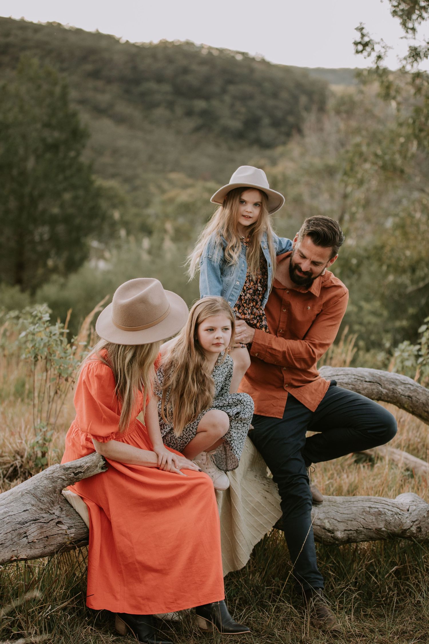 Family of four in autumn outfits sitting on fallen log in scenic outdoor setting.
