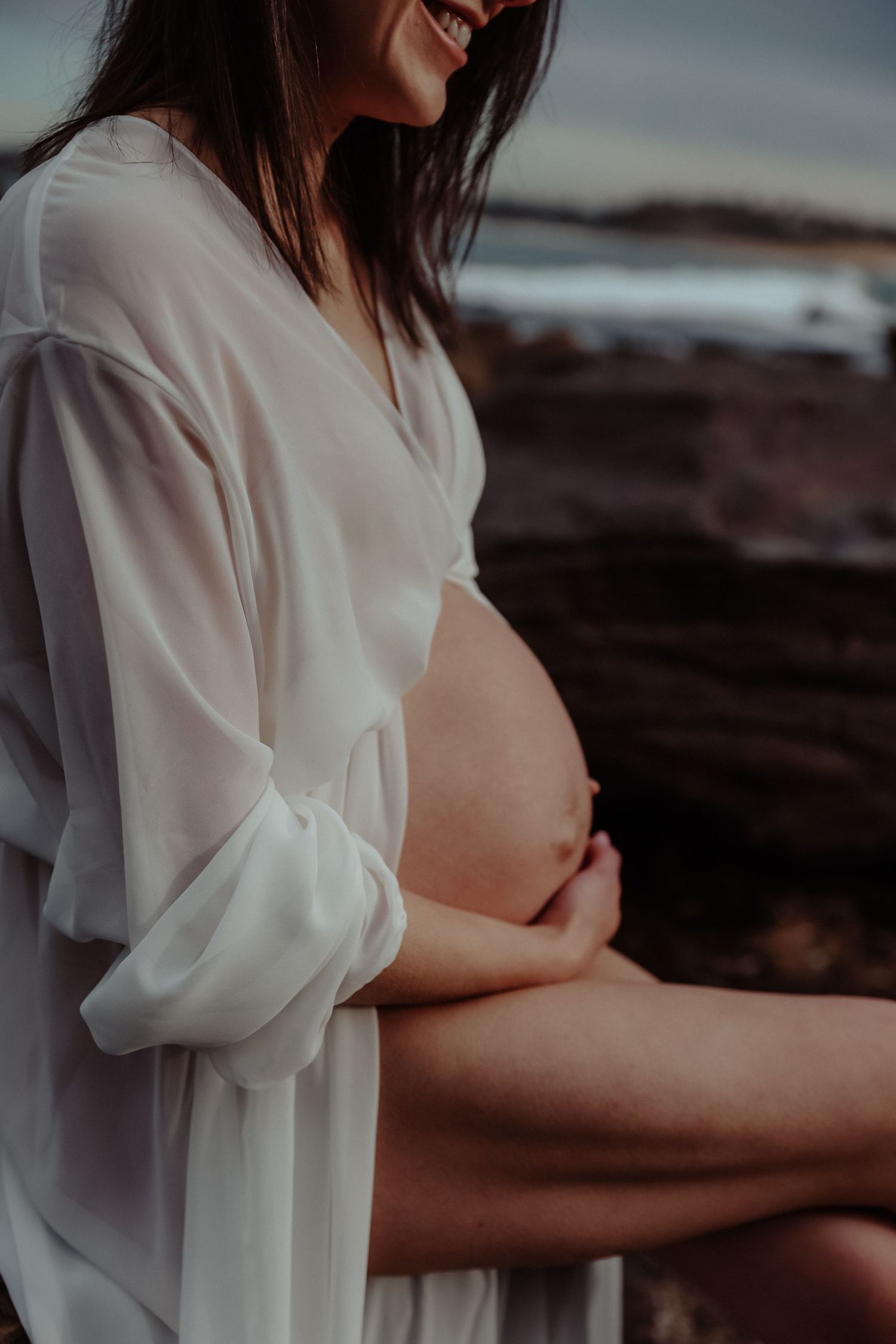 Close-up of pregnant woman in white sheer shirt cradling baby bump outdoors.
