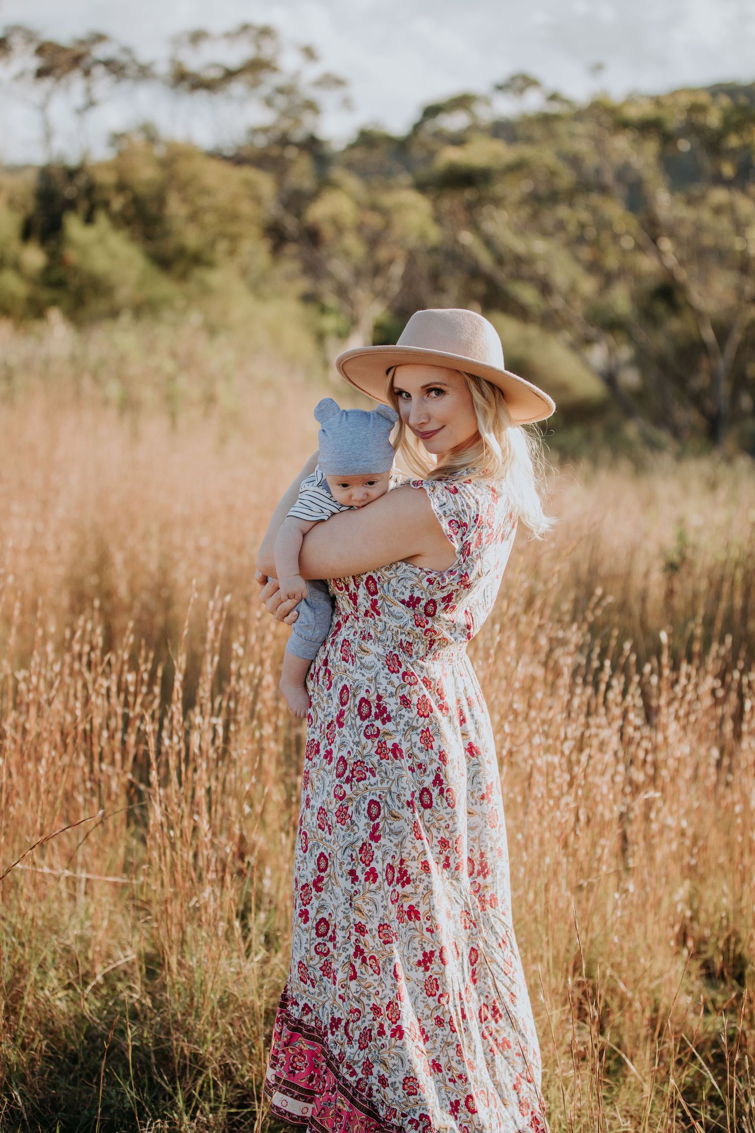 Blonde mother in floral maxi dress and hat holds baby in golden grass field.
