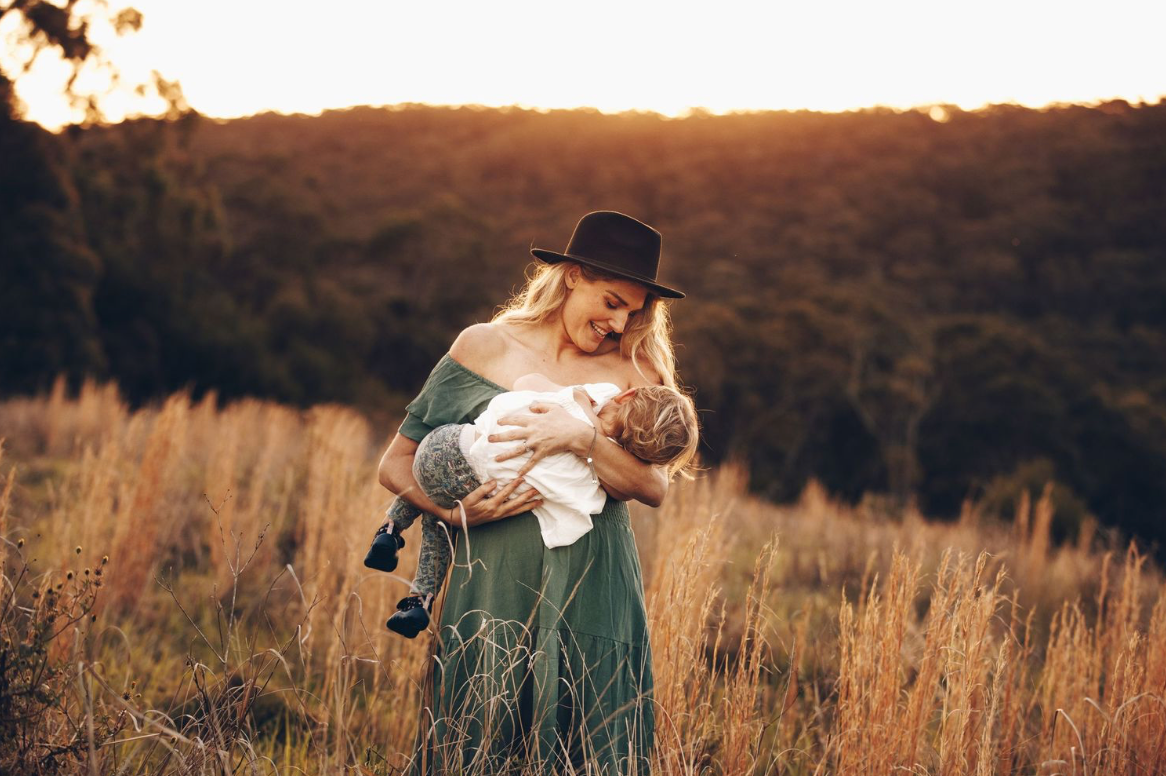 Mother in green dress and black hat cradling newborn baby in glowing sunset meadow.