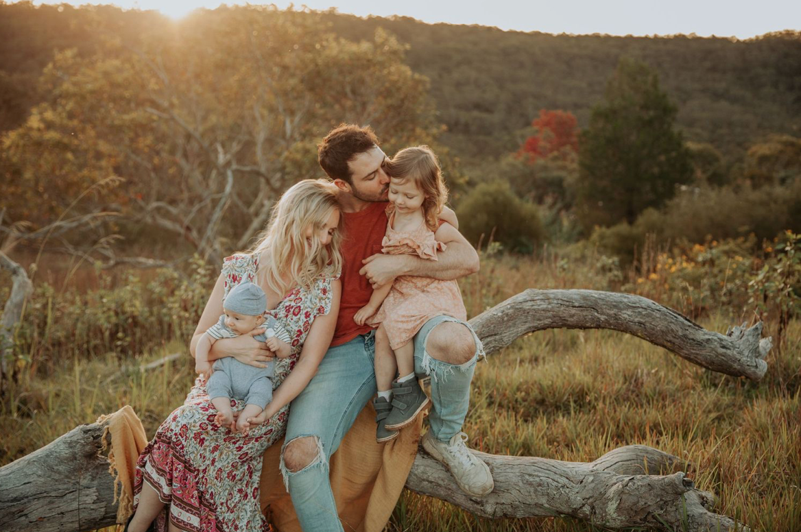 Family of four sitting on a fallen log in a golden sunlit autumn meadow.