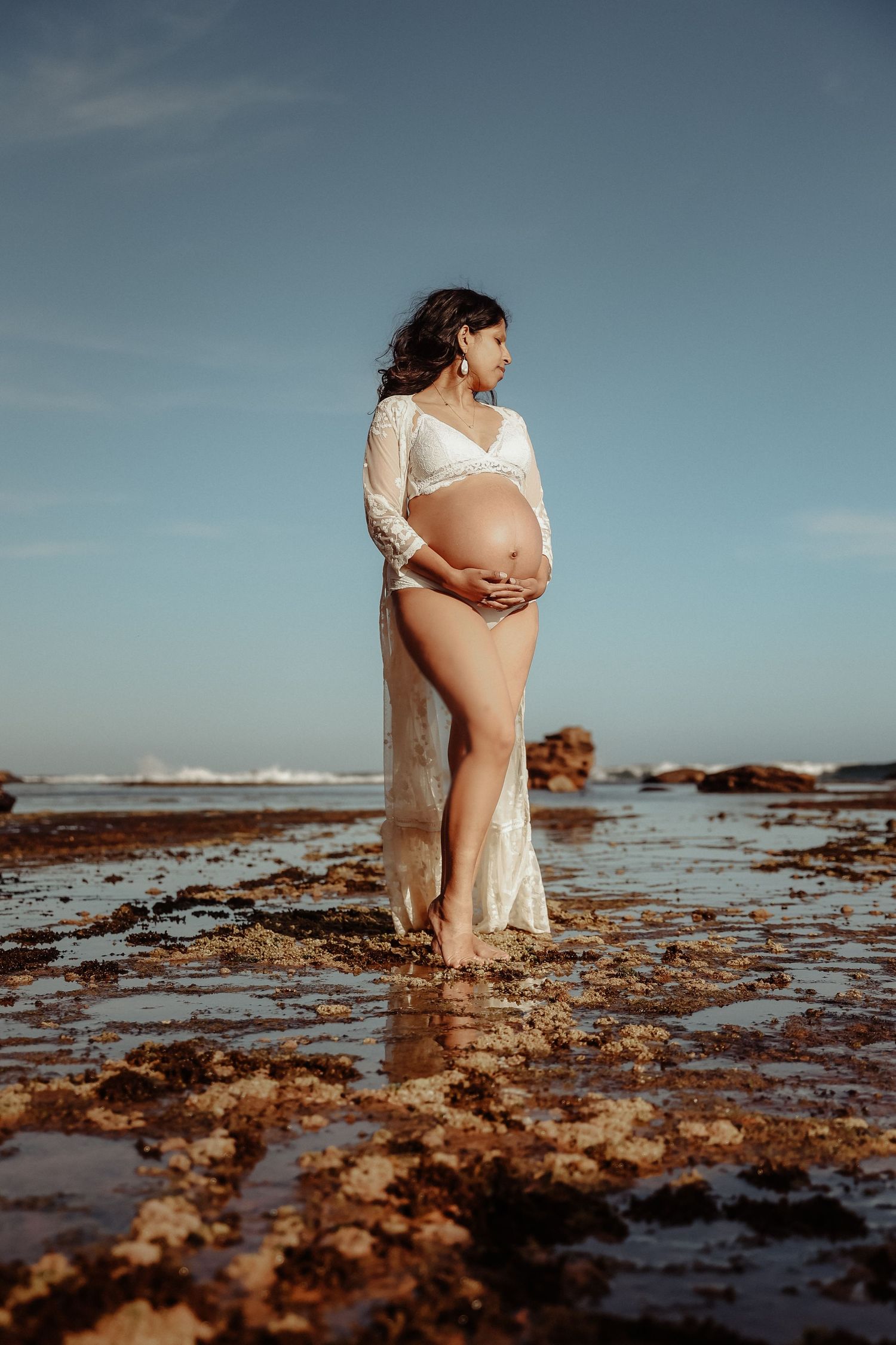 Pregnant woman in white lace dress cradling baby bump on rocky beach at sunset.