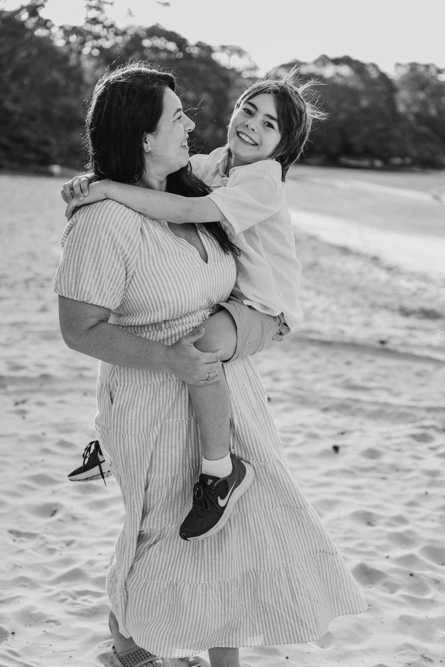Woman in striped dress holding smiling child on sandy beach in black and white photo.