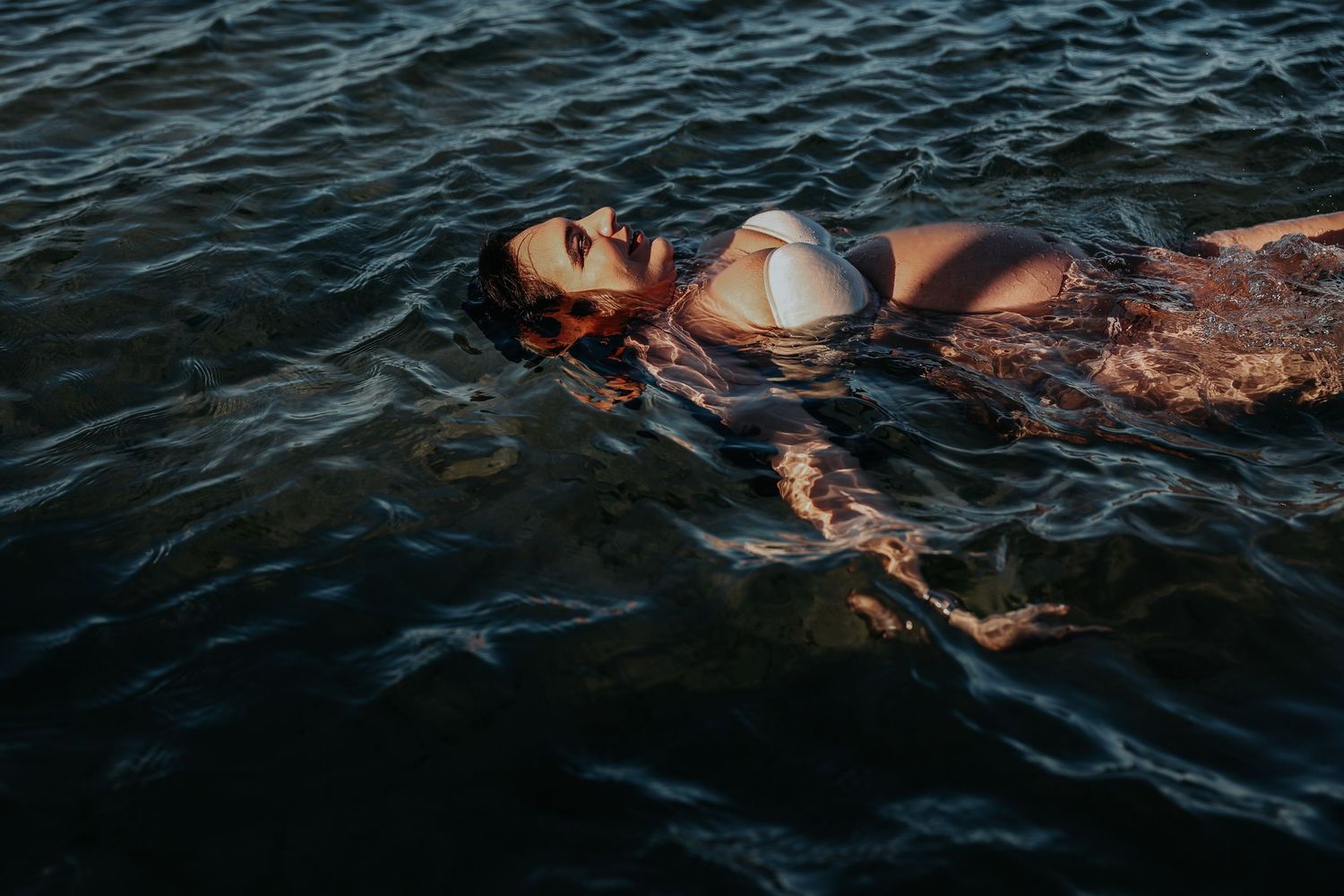 Pregnant woman floating peacefully on her back in dark ocean water during golden hour light.