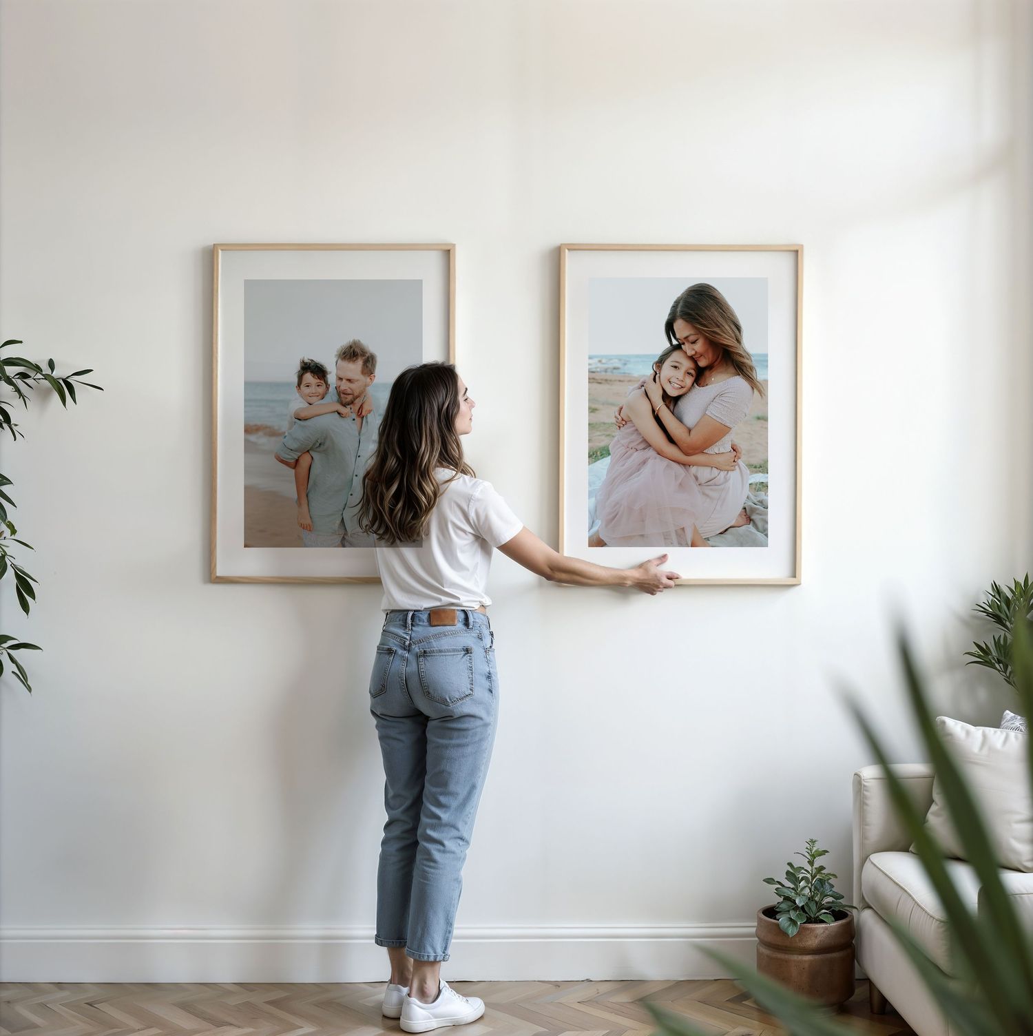 A woman hangs two large framed family beach photos side by side on a white interior wall.