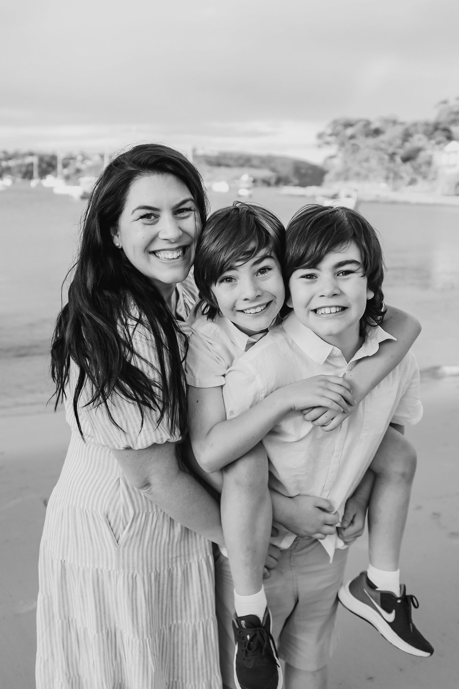 Black and white photo of a smiling mother hugging her two young sons on a beach.