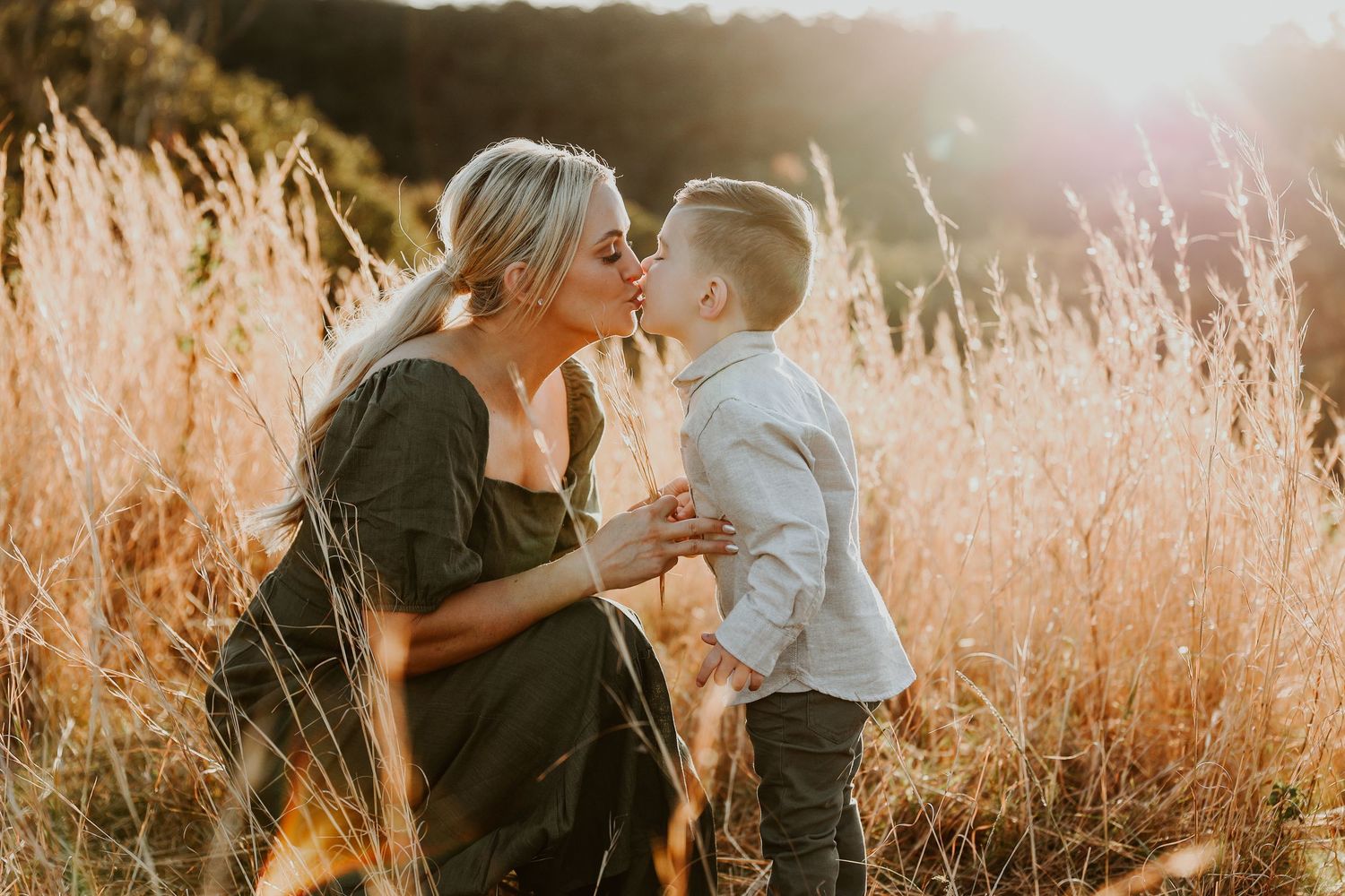 A pregnant woman kisses her young son in a golden sunlit field of tall grass during a maternity photo session.