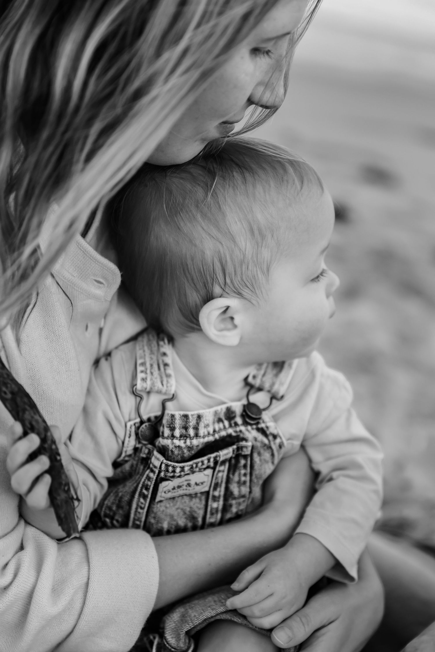 Black and white photo of a mother tenderly holding her baby in overalls outdoors.