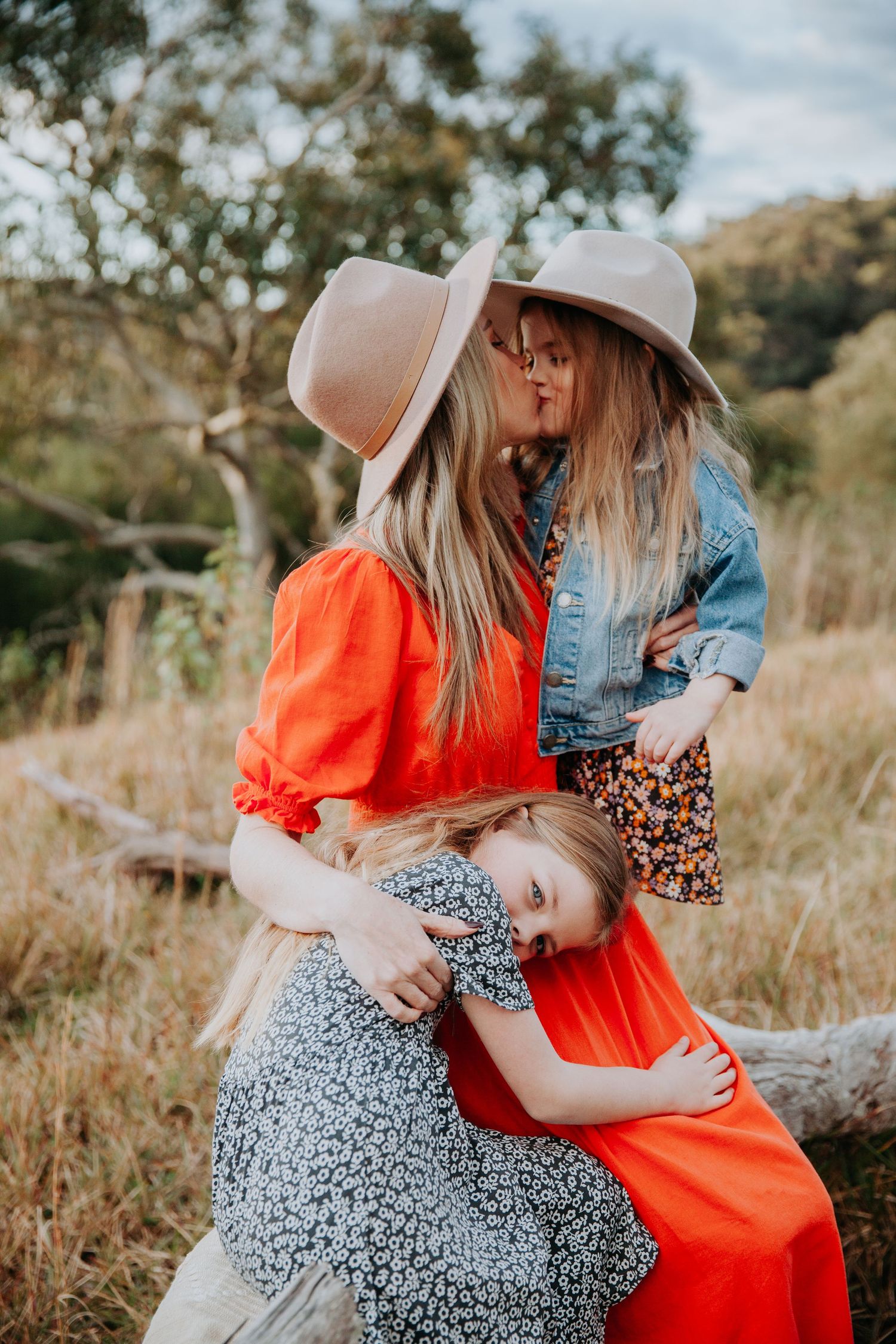 Mother and two daughters sharing a kiss outdoors wearing matching beige hats.