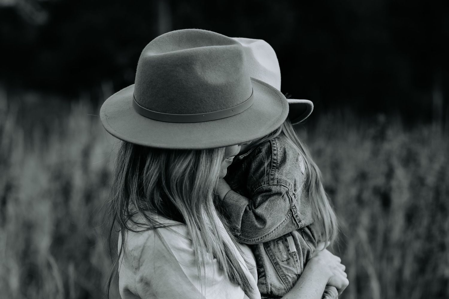 Black and white photo of a woman wearing a fedora hat hugging a child in a grassy field.