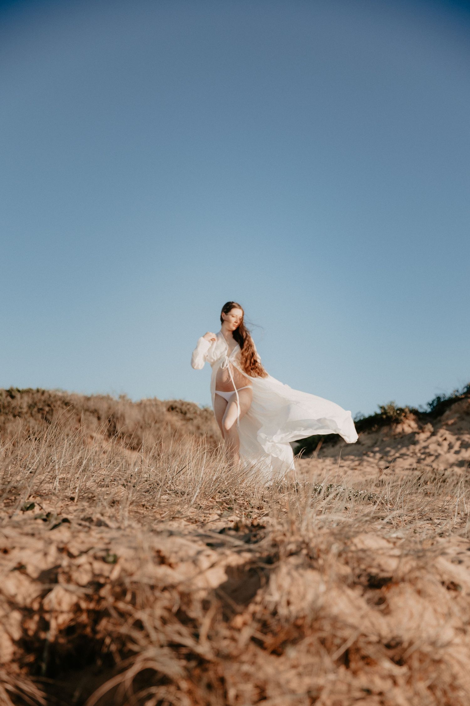 Pregnant woman in flowing white sheer robe standing on sandy dunes under clear blue sky.