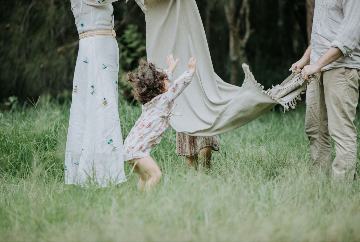 A young child plays in a grassy field while parents hold a flowing white blanket.