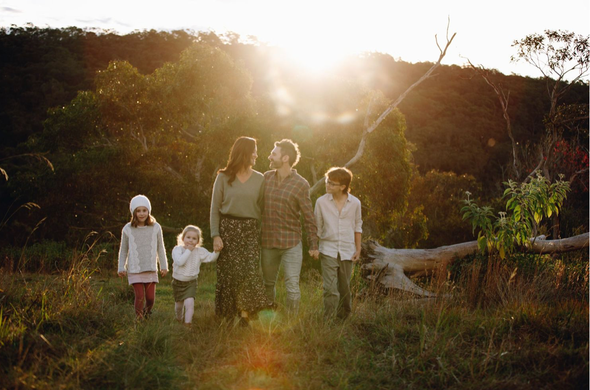 Family of five walking hand in hand through a sunlit meadow with golden hour light behind them.