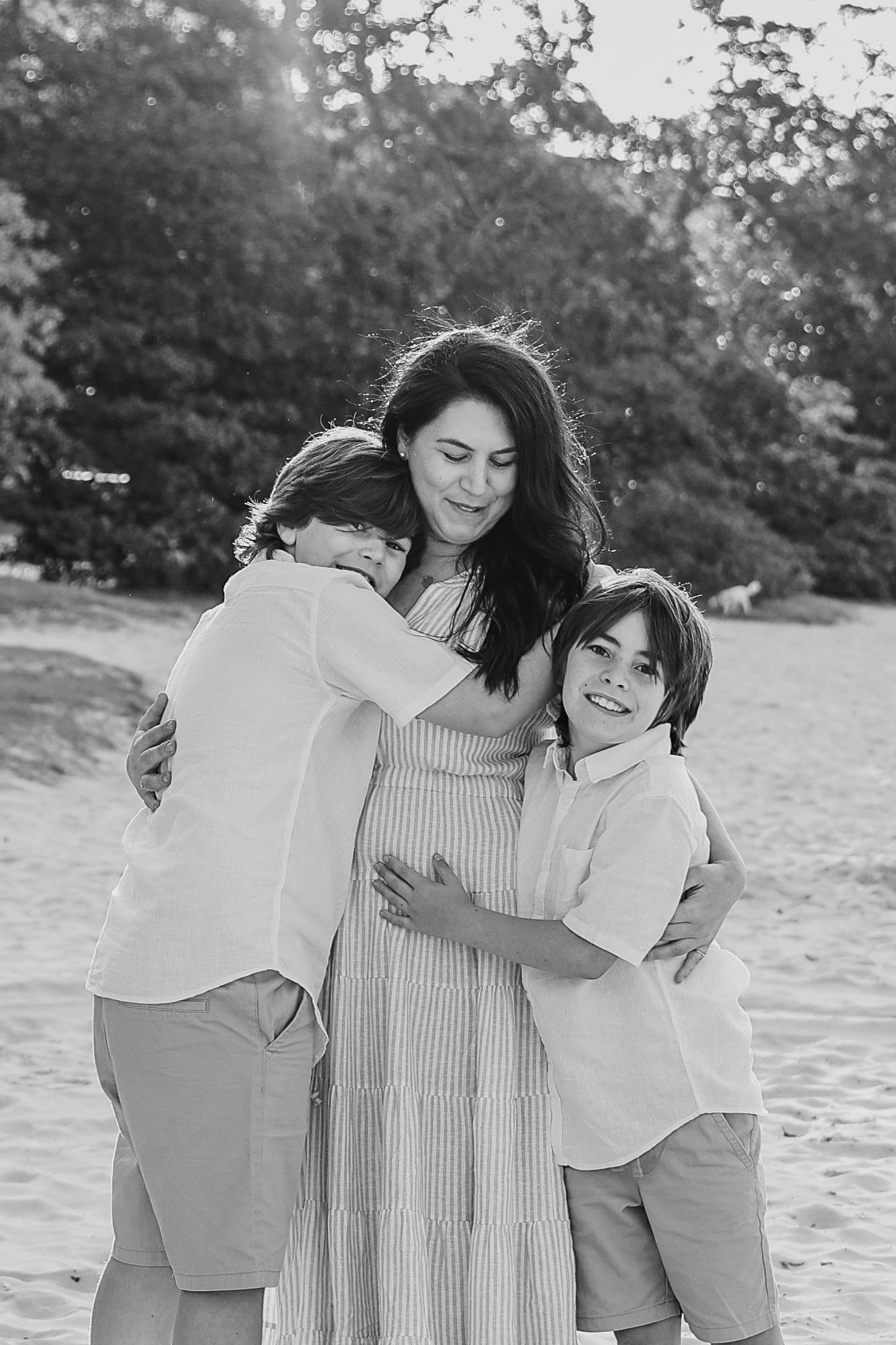 Black and white photo of a pregnant woman on a beach being hugged by two young boys in white shirts.