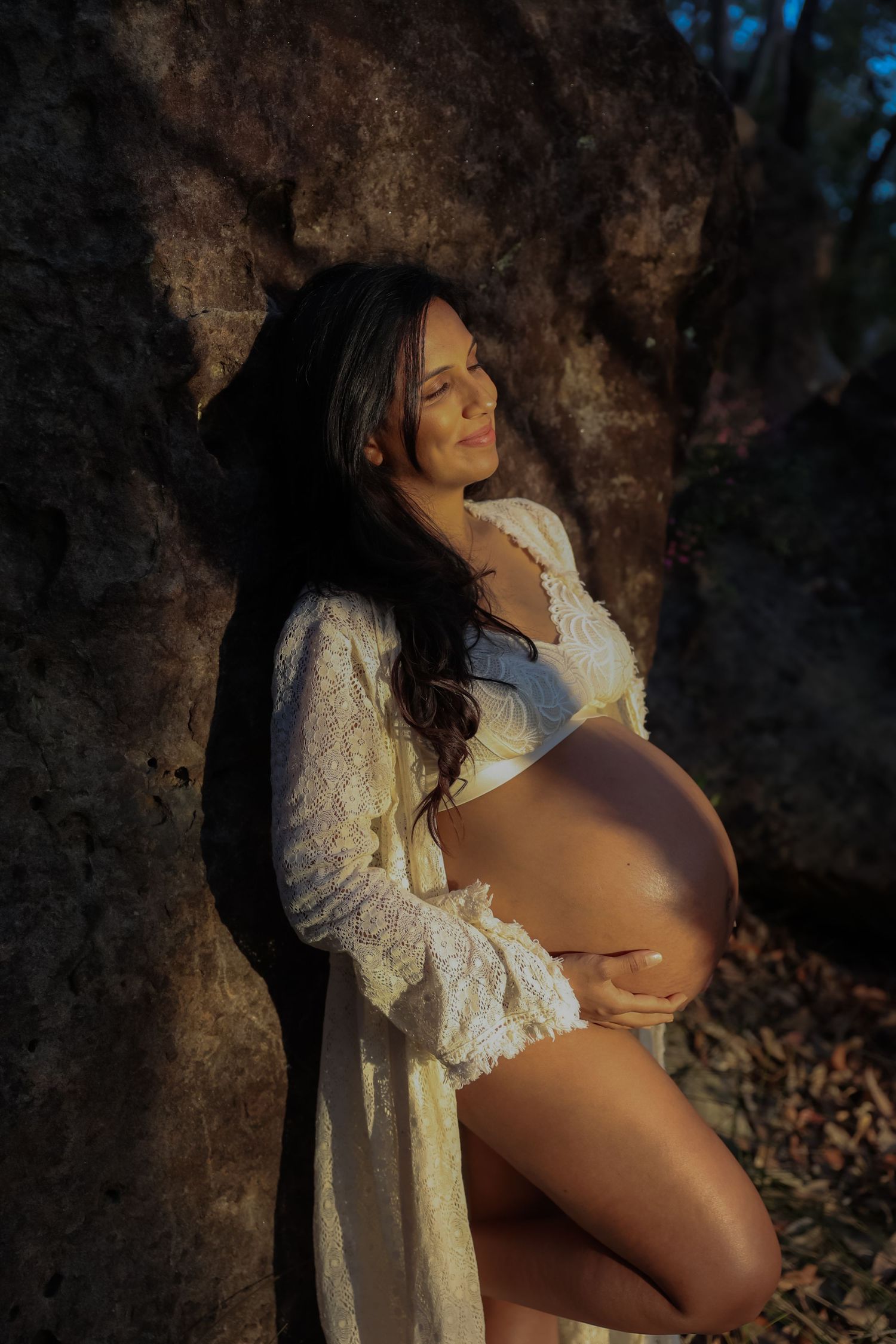 Pregnant woman in white lace robe cradling her baby bump against a rocky backdrop in warm sunlight.