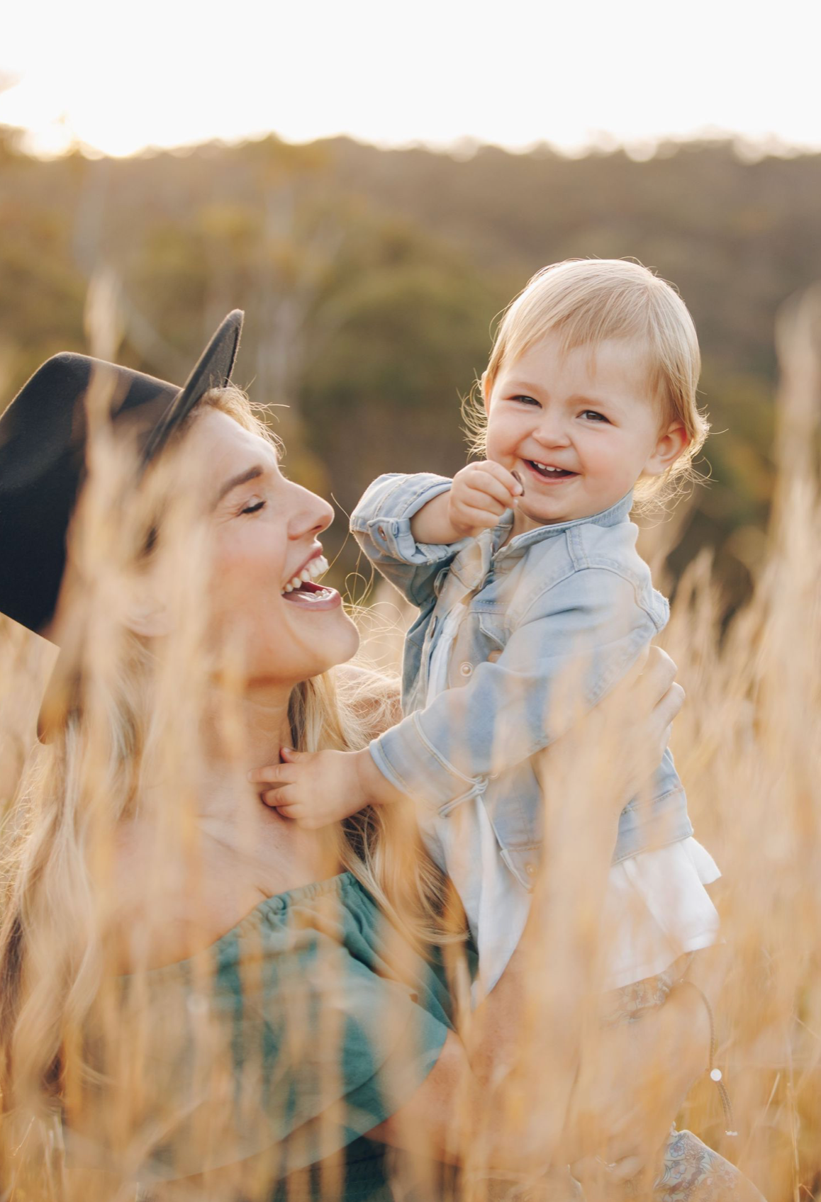 A joyful mother in a hat lifts her laughing baby in a golden wheat field at sunset.
