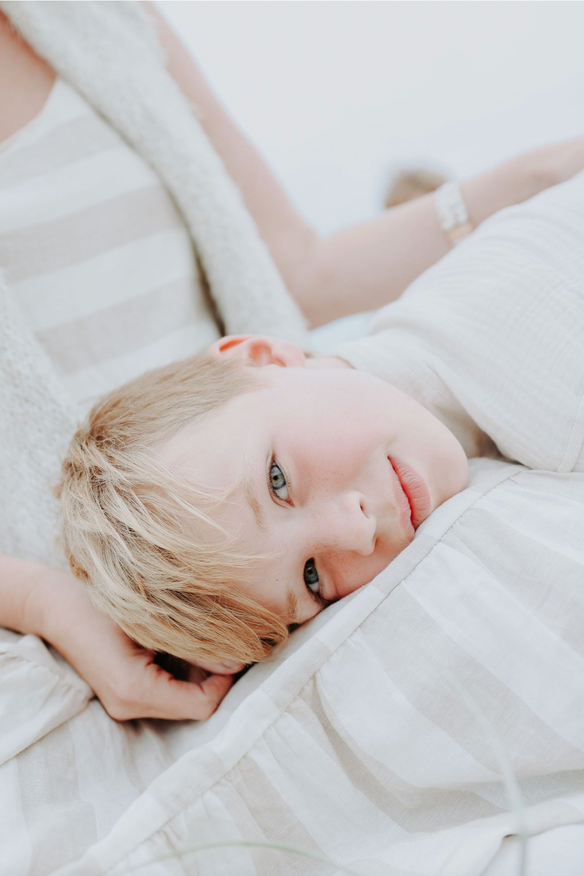 Young blonde child with blue eyes resting head on white bedding, looking up at camera softly.