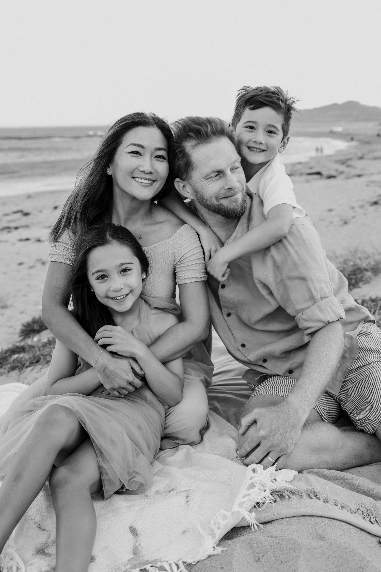 Black and white portrait of a loving family of four sitting together on a blanket at the beach.