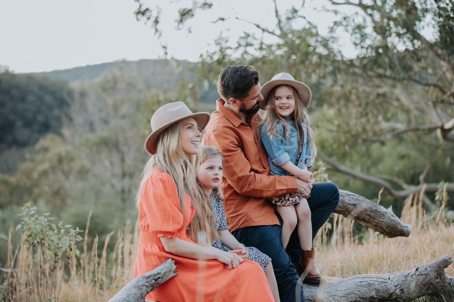 Family in coordinating earth tones relaxing on fallen log in natural hillside setting.