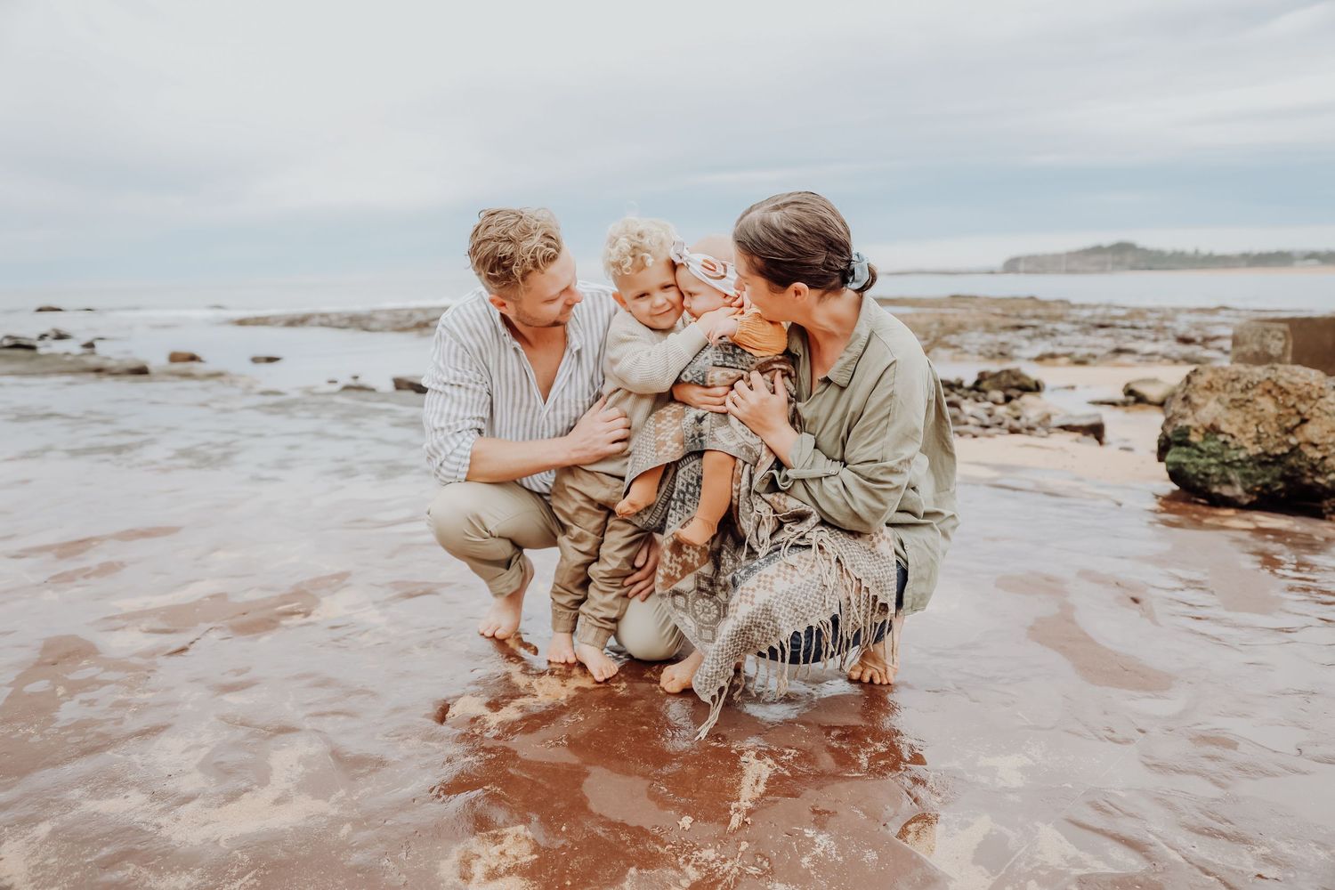 Two parents crouching on a sandy beach with their young child, smiling and playing in shallow water.