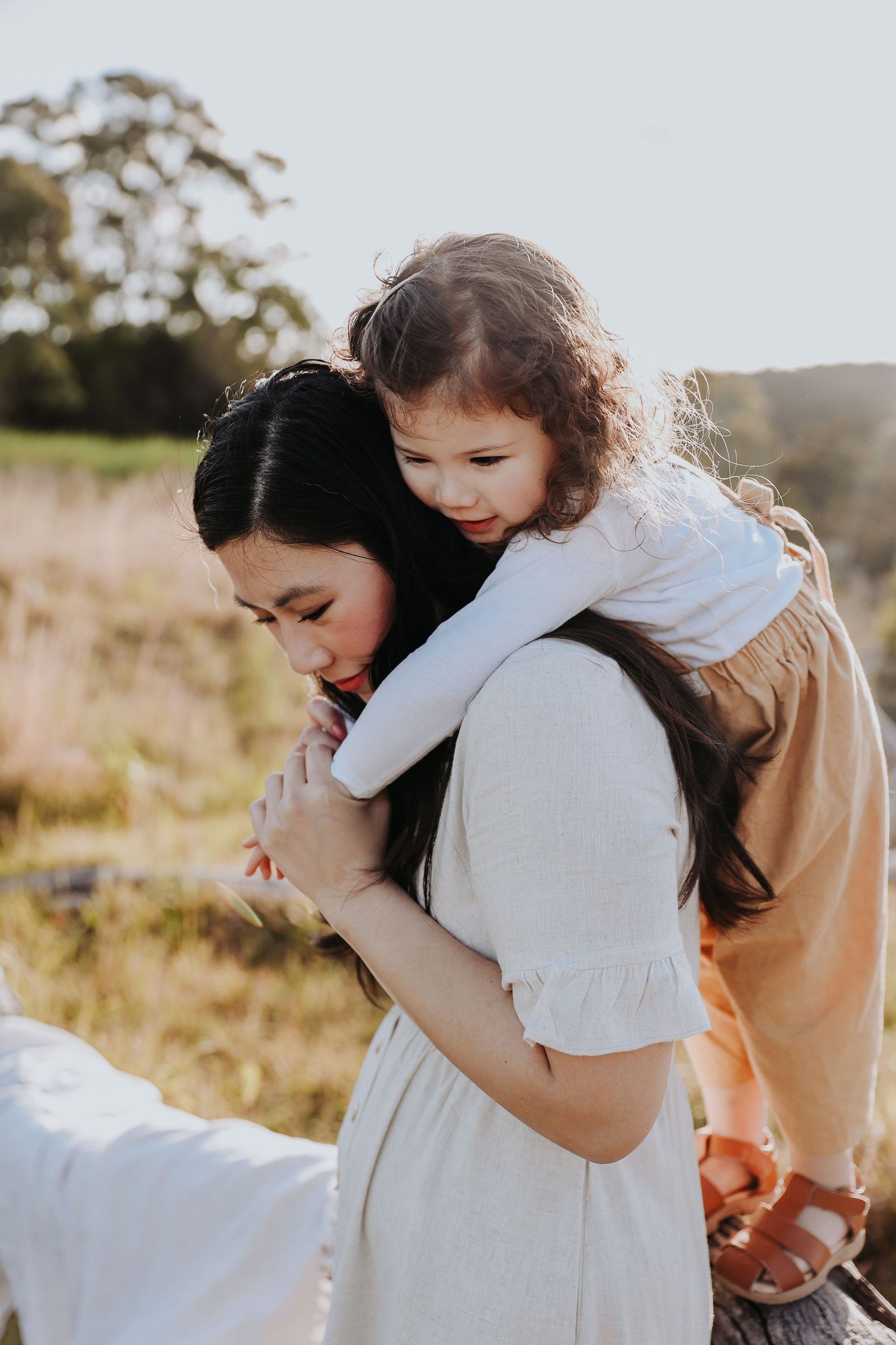 A young girl hugs her dark-haired pregnant mother from behind in a sunlit meadow.