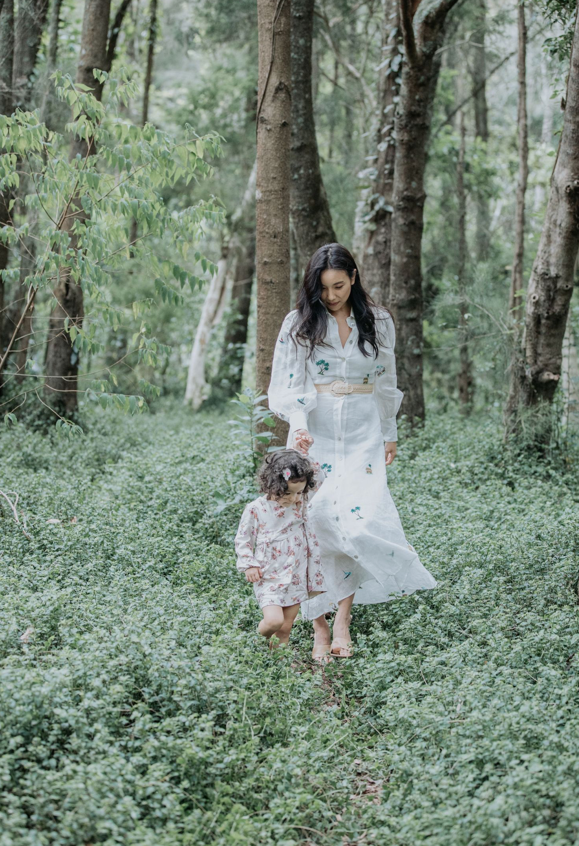 A mother and toddler walk hand in hand through a lush green forest setting.