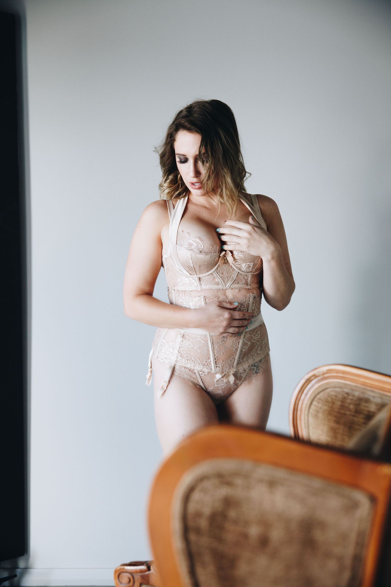 Woman in beige lace lingerie corset standing in a bright room beside a vintage chair.