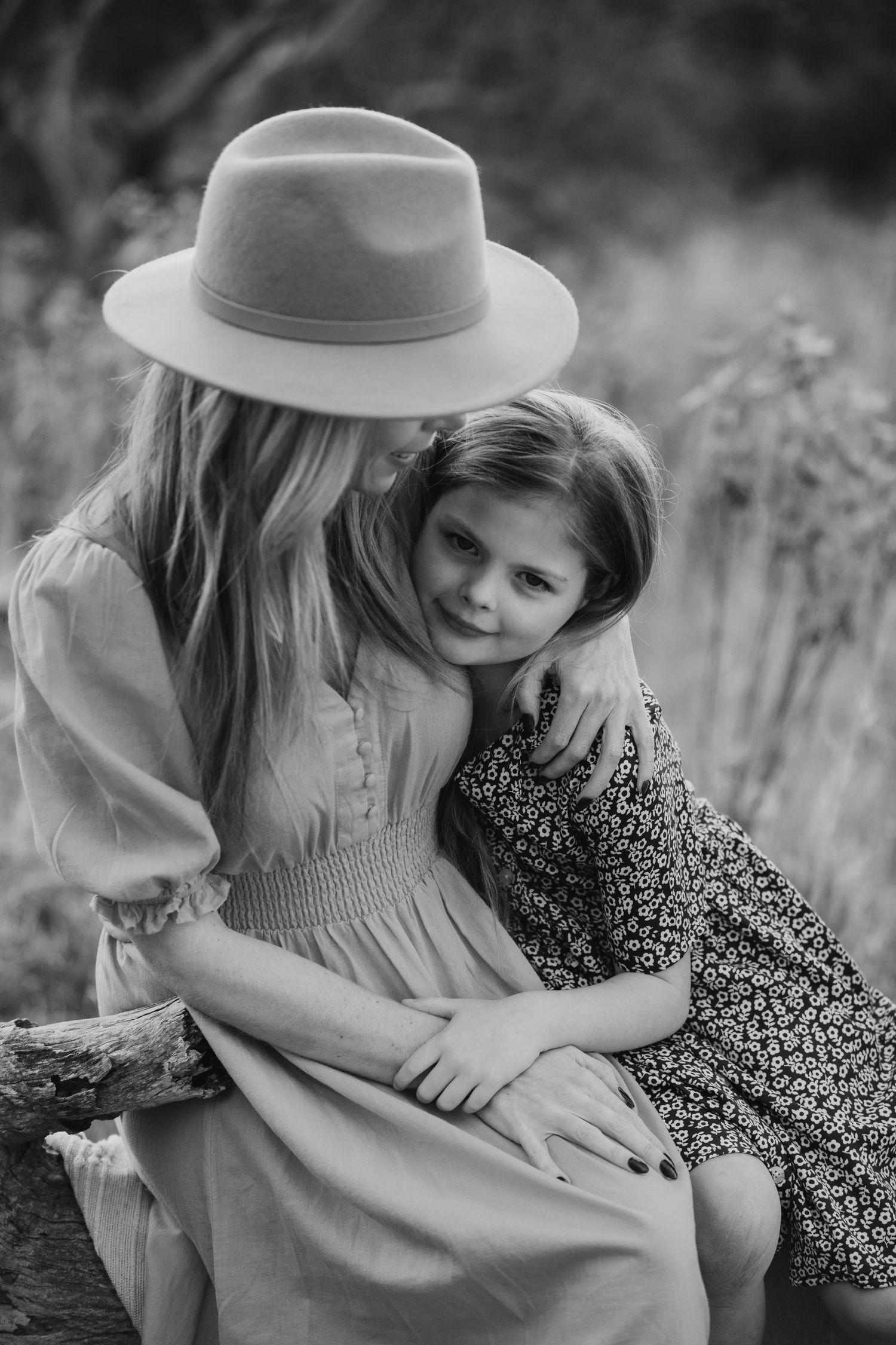 Black and white photo of a woman in a wide-brim hat hugging a young girl outdoors in a garden setting.