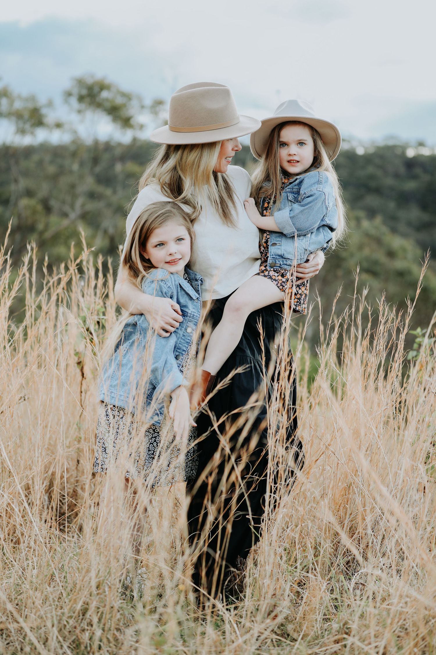 Mother in hat holds two young daughters in denim jackets amid golden tall grass field outdoors.