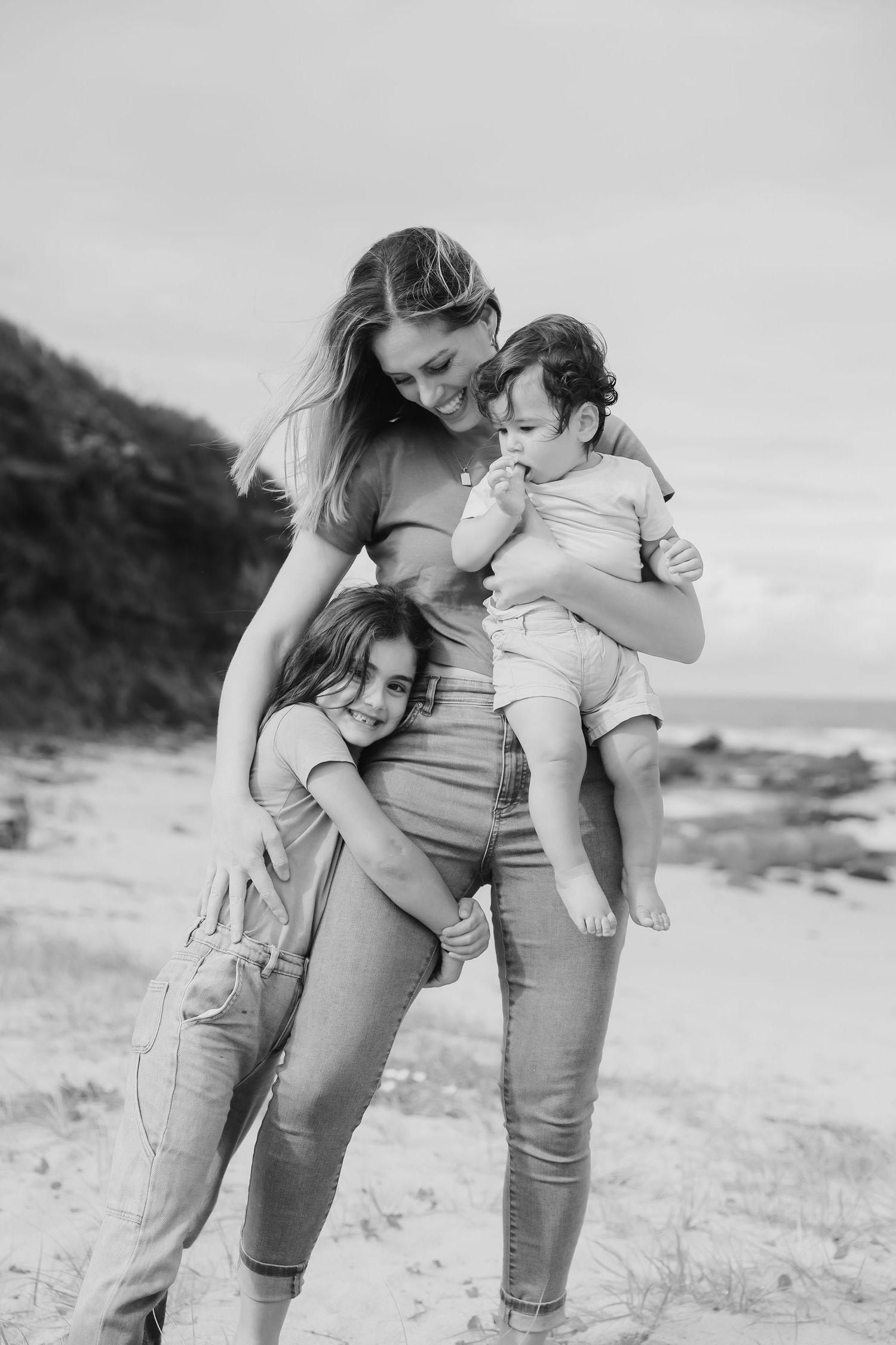 Mother smiling at baby on beach while older child hugs her waist, black and white family photo.
