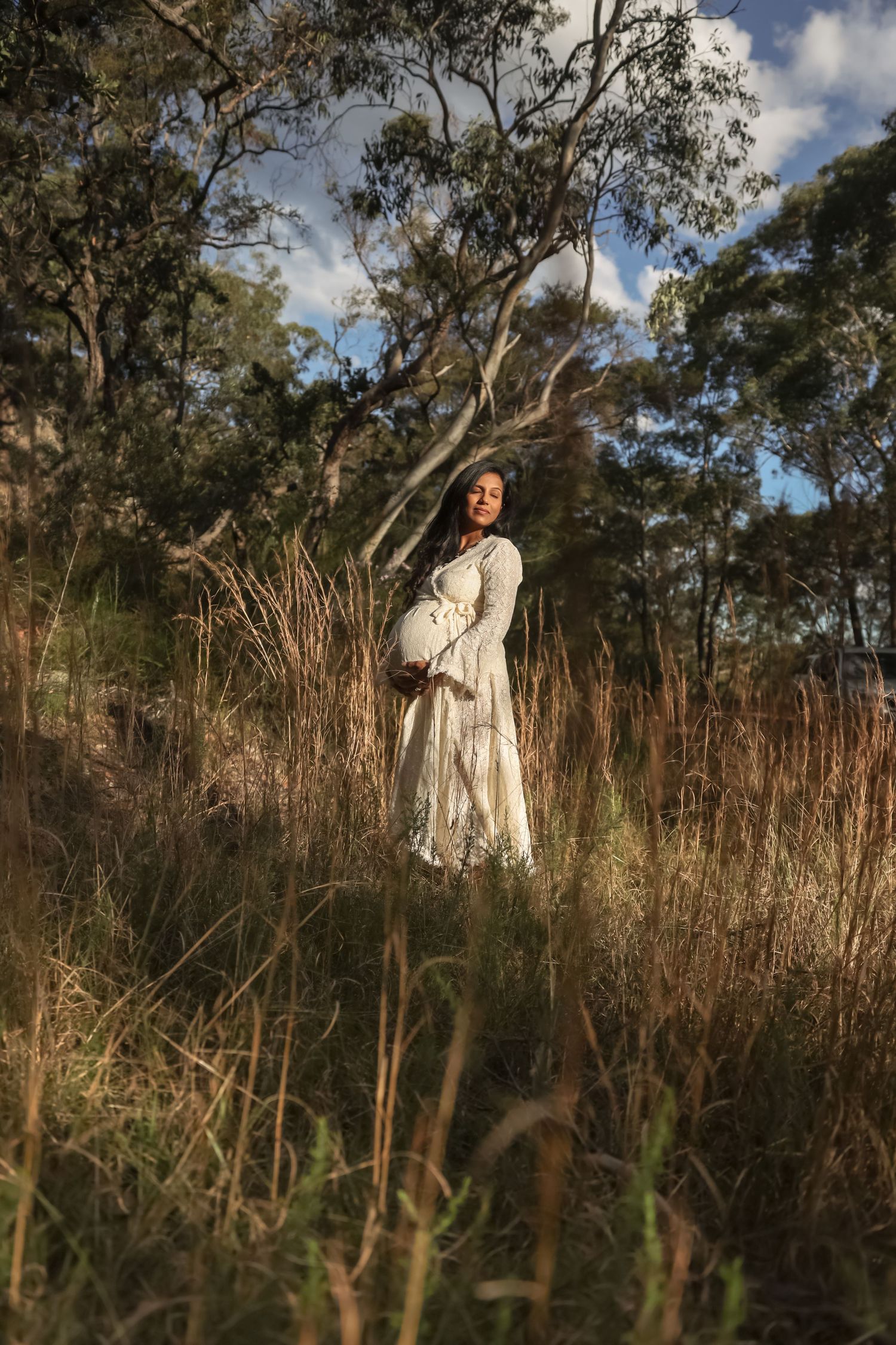 Pregnant woman in white dress standing in tall grass surrounded by eucalyptus trees in golden light.