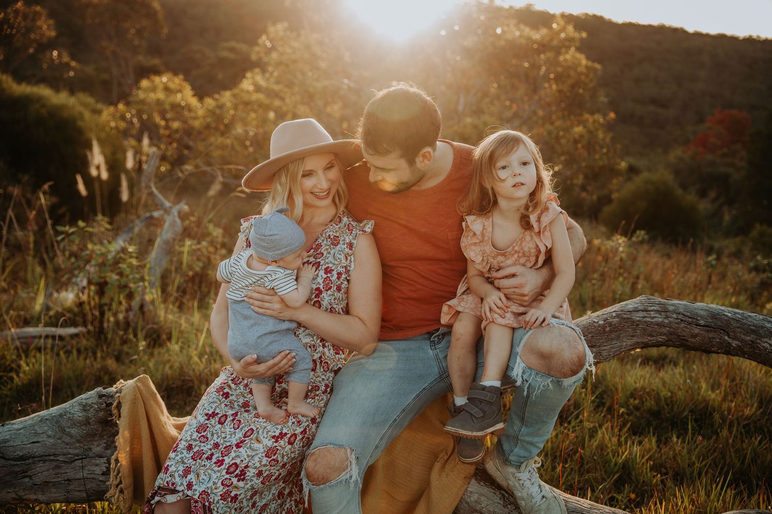 Family of four sitting on a log in golden sunset light, parents holding young children in a rustic outdoor setting.