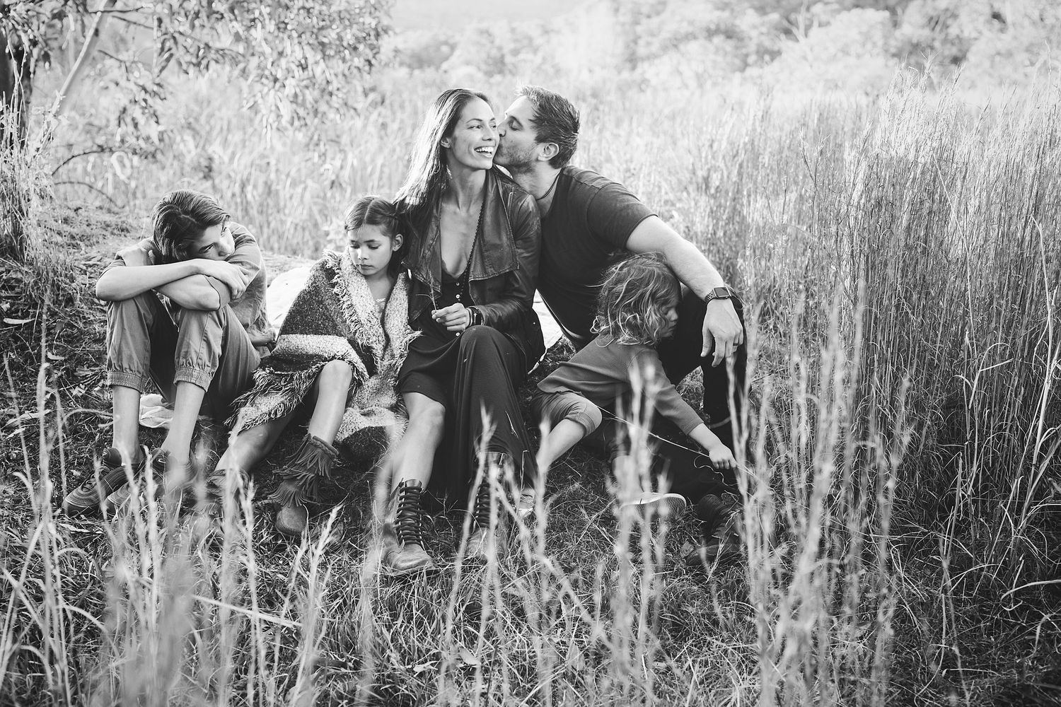 Black and white family photo of parents and children sitting together in tall grass field, sharing a joyful moment.