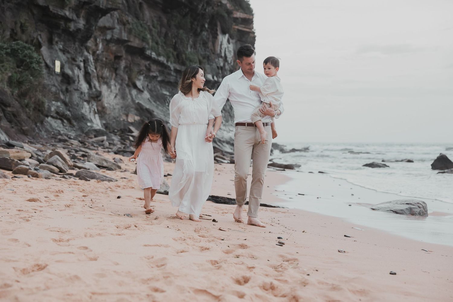 Family of four walking on a sandy beach with rocky cliffs, wearing white and neutral tones in a serene coastal setting.