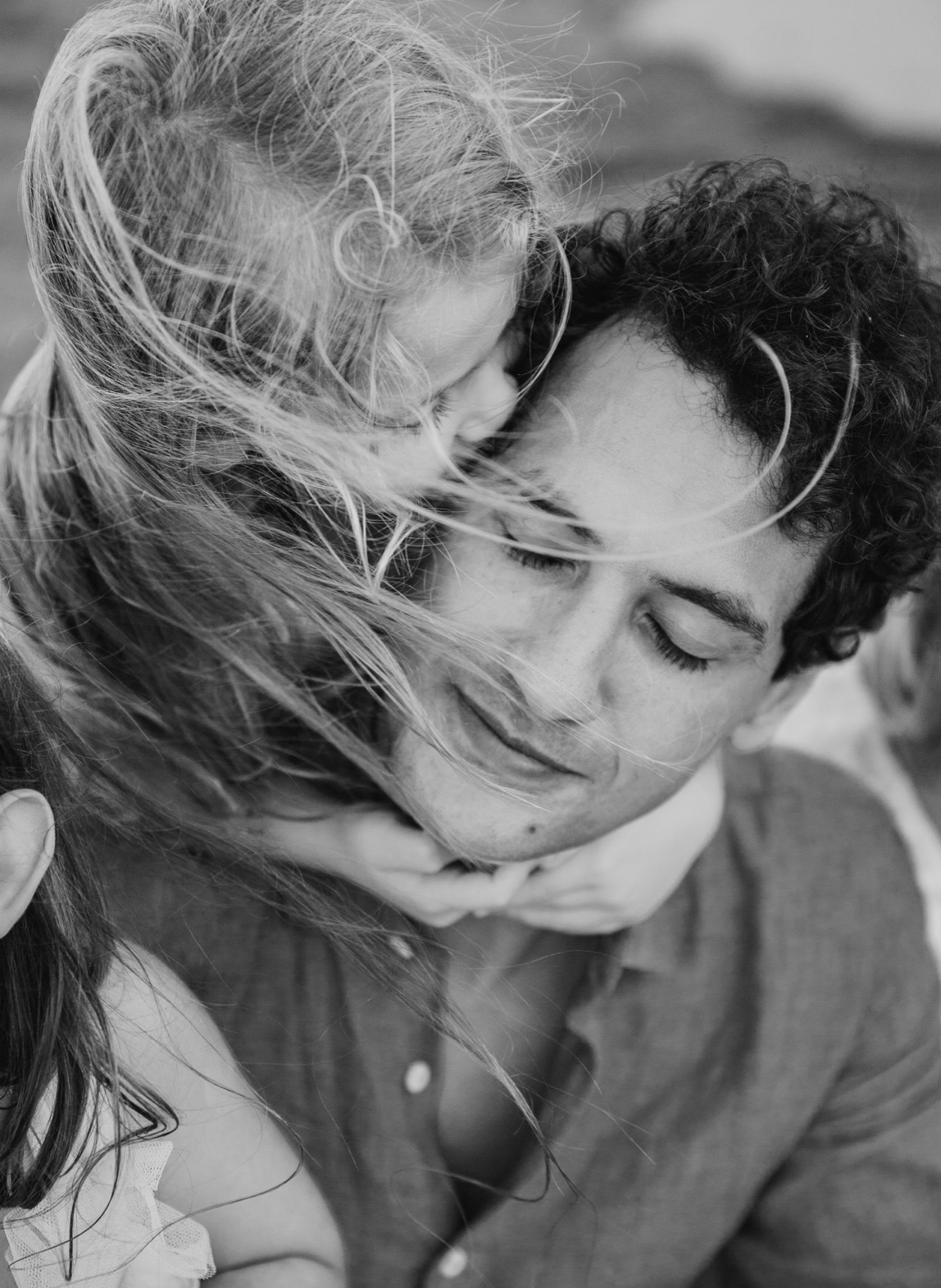 Black and white photo of a couple embracing outdoors, woman's hair blowing in the wind as man closes his eyes.