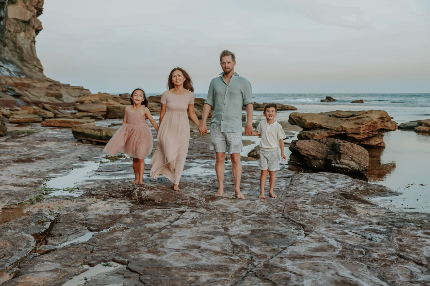 Family of four holding hands walking on rocky beach shoreline at dusk in casual attire.