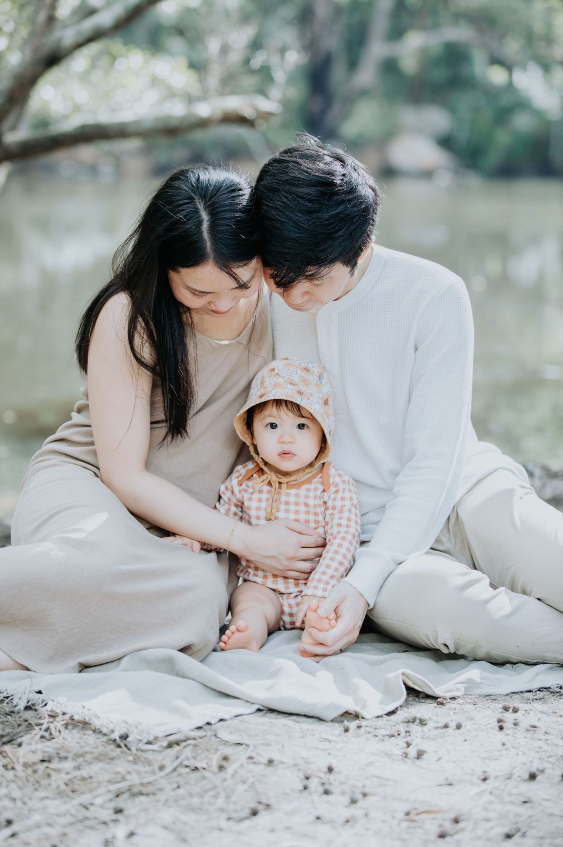 Young couple sitting outdoors with their baby on a blanket by a lake.